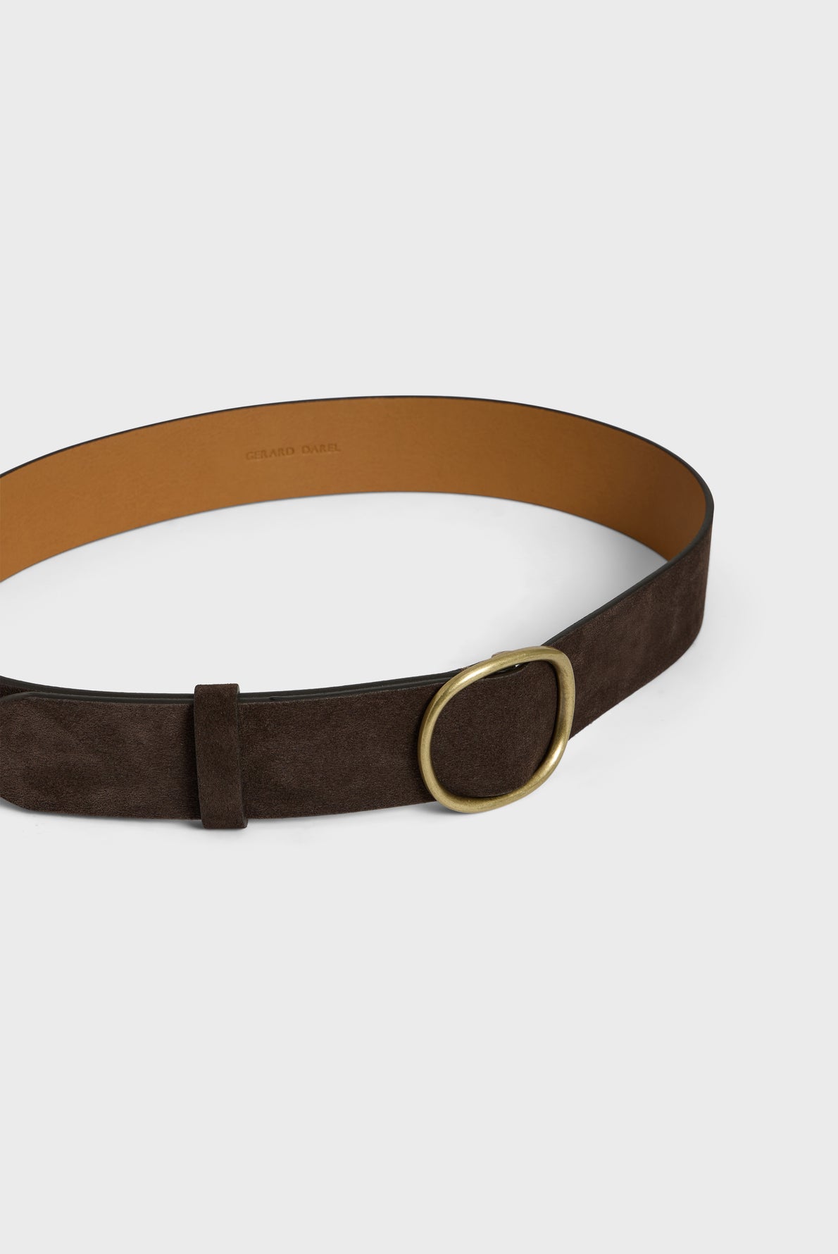 A dark brown cuir velours ceinture minimaliste with a gold circular buckle is lying flat on a light background. The inside of the belt is tan.