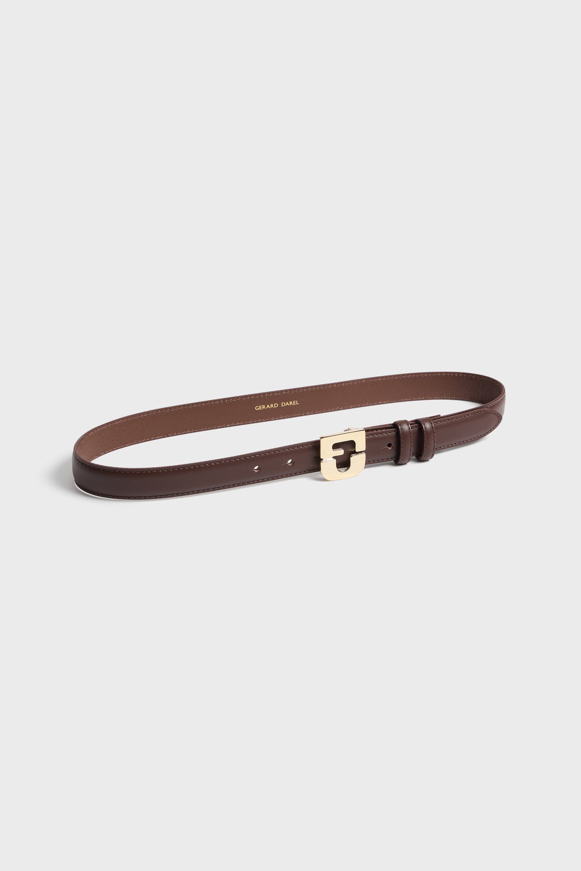 A brown leather belt with a gold buckle is laid flat on a light gray background. The interior of this ceinture élégante is lighter brown, and Gerard Darel is printed inside in gold lettering.