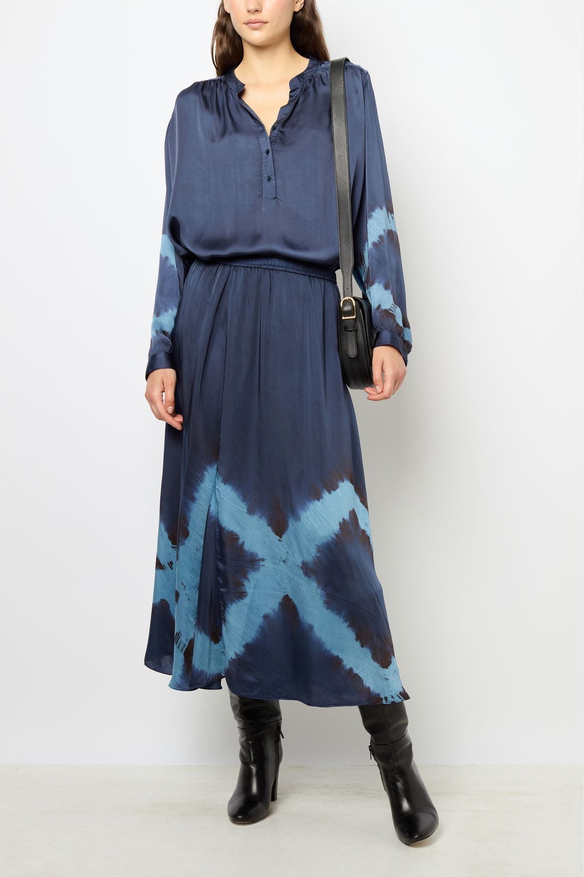 Woman wearing a navy blue tie and dye dress with long sleeves, a matching jupe midi, black knee-high boots, and a black shoulder bag, standing against a plain light background.