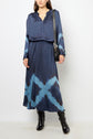Woman wearing a navy blue tie and dye dress with long sleeves, a matching jupe midi, black knee-high boots, and a black shoulder bag, standing against a plain light background.