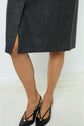 A person wearing a grey motif Prince-de-galles plaid skirt with a small slit and black slingback shoes stands on a light-colored floor.