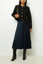 A woman wearing a black jacket with gold buttons, a navy blue jupe plissée with an aspect satiné, black knee-high boots, and carrying a black shoulder bag, standing against a plain light background.