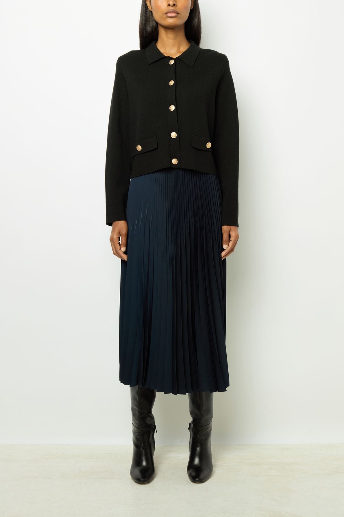 A person wearing a black buttoned jacket, a navy jupe plissée midi with an aspect satiné, and black knee-high boots stands against a plain white background.