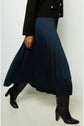 A person in a black jacket and pleated navy skirt walks, wearing knee-high black heeled boots. The jupe plissée catches the light with an elegant aspect satiné, while only the lower body and part of the jacket are visible against a plain background.