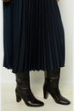A person wearing a dark jupe midi plissée and black leather heeled knee-high boots stands on a light floor.
