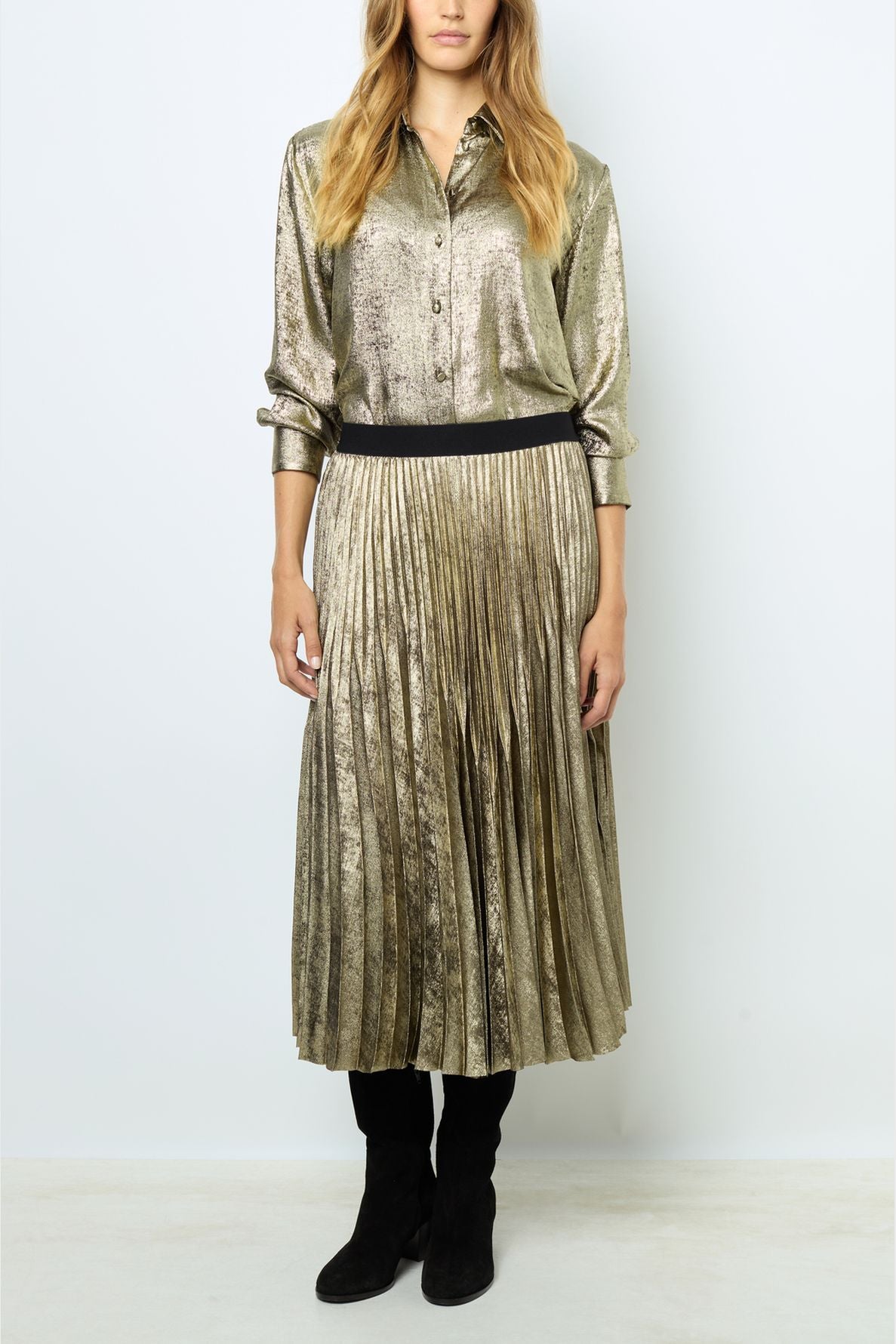 Woman wearing a shiny gold button-up blouse and a matching gold pleated skirt, with black boots. She stands against a plain light background, with her hands resting naturally by her sides.