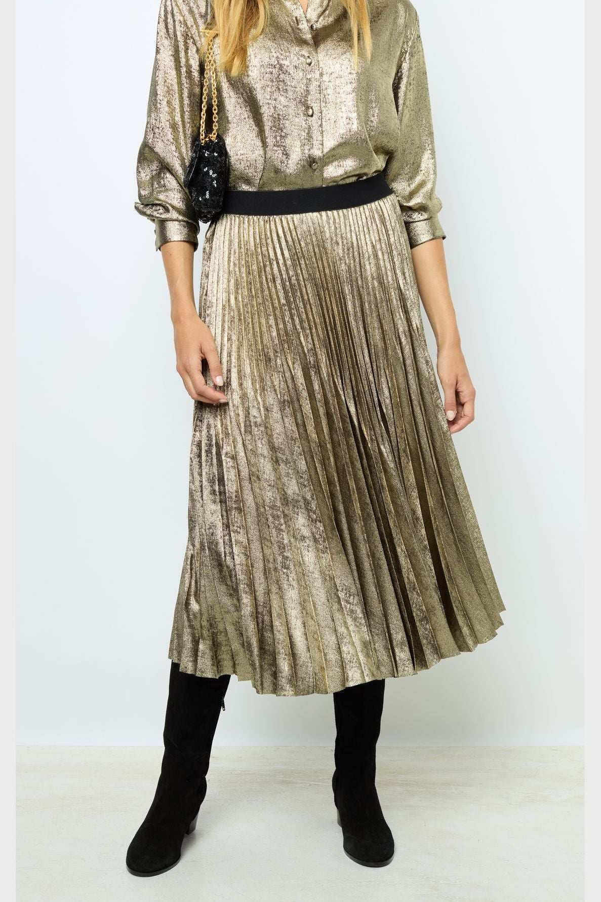 A person wearing a shiny gold pleated skirt, matching gold blouse, black knee-high boots, and holding a small black purse with a gold chain. The head is not visible in the image.