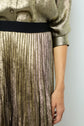 A person wearing a shiny, metallic gold blouse and a pleated gold and pink metallic skirt, with their right arm relaxed at their side, shown from the shoulder to knee.
