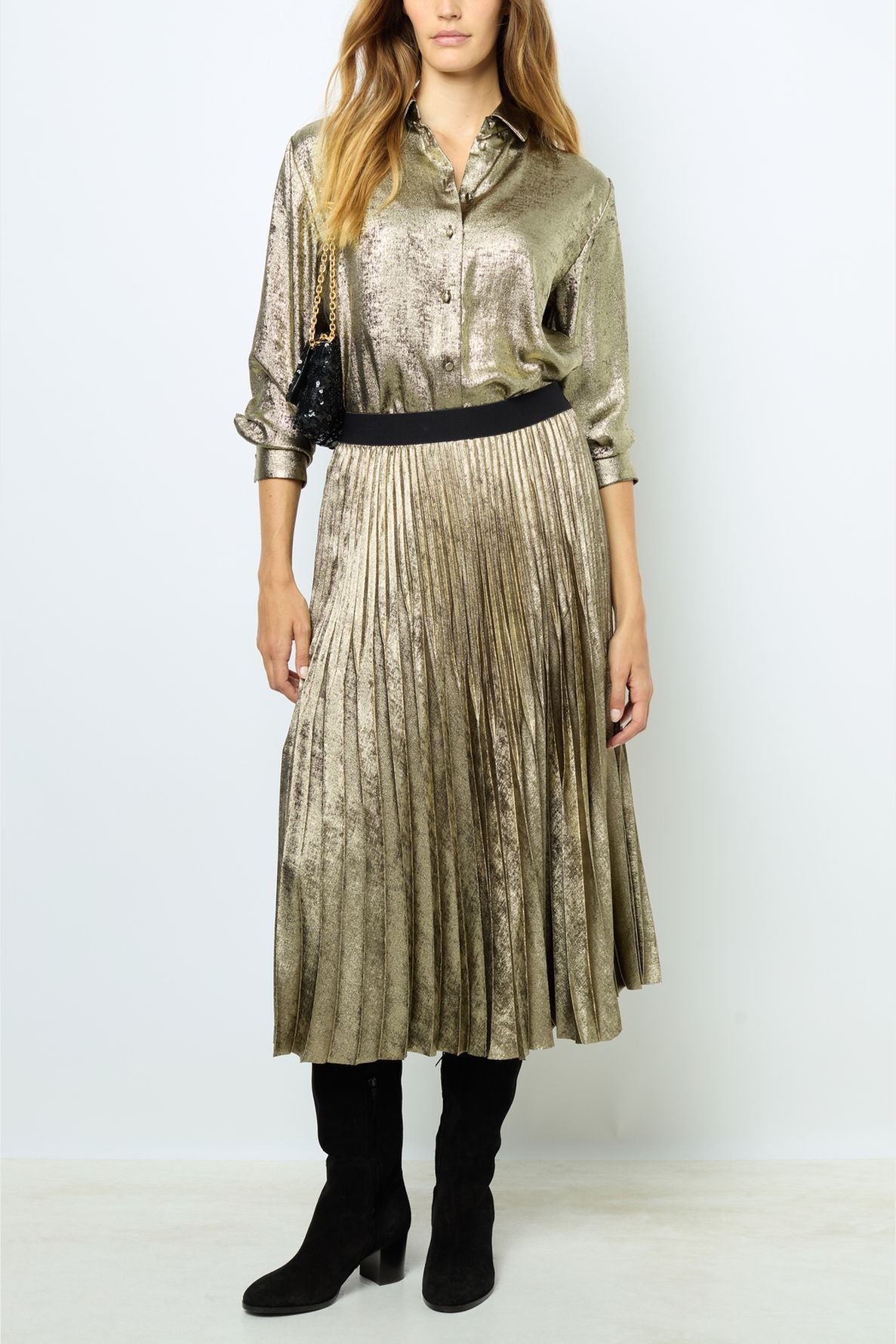 A woman wearing a shiny gold blouse and matching pleated skirt, black belt, black knee-high boots, holding a small black purse. The background is plain white.
