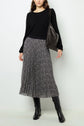 Woman wearing a black sweater, a black-and-white pleated midi skirt, black knee-high boots, and carrying a dark shoulder bag, standing against a plain light background.