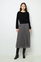 Woman standing against a white wall, wearing a black long-sleeve top, a black and white pleated skirt with a subtle pattern, and black knee-high boots.
