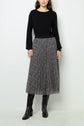 A person wearing a black long-sleeve sweater, a black-and-white pleated midi skirt with a small dot pattern, and black knee-high boots stands against a plain light background.
