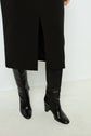 A person wearing knee-high black leather boots and a black skirt with a front slit stands on a light-colored floor against a white wall.