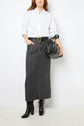 Woman wearing a white button-down shirt tucked into a long jupe midi en denim noir, cinched with a black belt and finished with black boots and a sleek black handbag. The jupe fendue adds a modern touch as she stands against a plain white background.