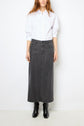 Person wearing a white button-up shirt tucked into a long, dark gray jupe midi en denim souple with a subtle jupe fendue, paired with black boots, standing against a plain white background.