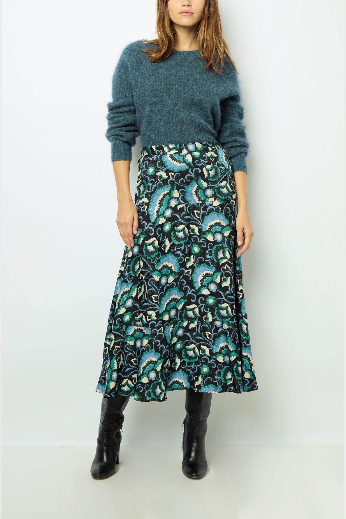 A woman wearing a teal fuzzy sweater, a long patterned skirt with blue, green, and white floral designs, and black knee-high boots stands against a plain light background.