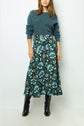 A woman wearing a teal fuzzy sweater, a long patterned skirt with blue, green, and white floral designs, and black knee-high boots stands against a plain light background.