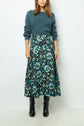 Woman wearing a fuzzy teal sweater, a long patterned skirt with green, teal, and cream designs, and black knee-high boots, standing against a plain light-colored background.