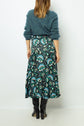 A woman stands facing away, wearing a fuzzy teal sweater, a long teal and black patterned skirt, and black knee-high boots against a plain white background.
