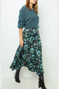 Woman in a teal fuzzy sweater, patterned teal and black midi skirt, and black knee-high boots stands against a plain white background. Her face is partially out of frame.