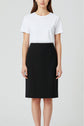 A woman stands facing forward, wearing a plain white short-sleeve t-shirt tucked into a jupe noire mi-longue, styled in a classic pencil skirt silhouette. Her hair is down and her hands rest at her sides against a plain, light background.
