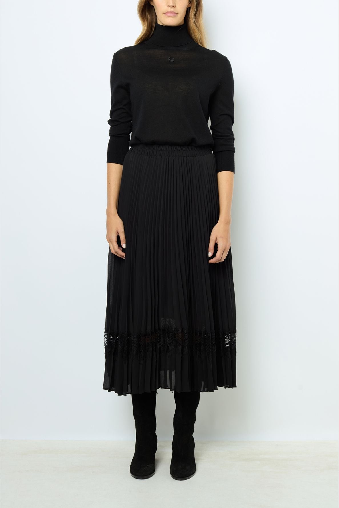 A person wearing a black turtleneck sweater, a long jupe plissée noire with a bande de dentelle at the hem, and black boots stands against a plain white background.