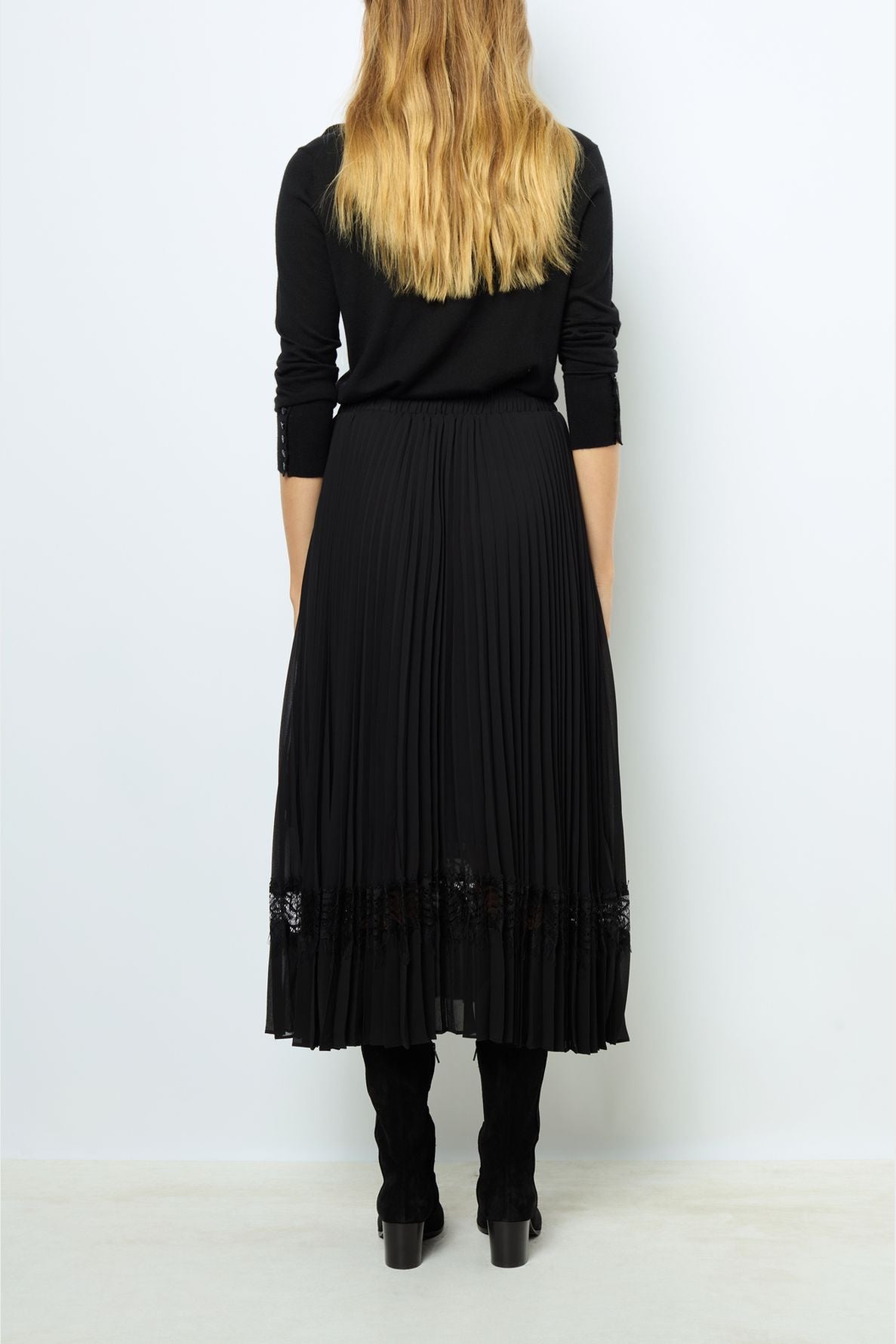 A woman with long, wavy blonde hair stands facing away, wearing a black long-sleeve top and a jupe midi plissée adorned with a delicate bande de dentelle at the hem, paired elegantly with black boots.