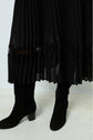 A person wearing a jupe midi noire plissée with a bande de dentelle near the hem and black heeled suede boots, standing on a light-colored floor.