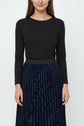 A woman wearing a long-sleeved black top and a dark blue jupe plissée midi skirt stands against a plain white background. Her face is partially visible, and her arms are relaxed at her sides.