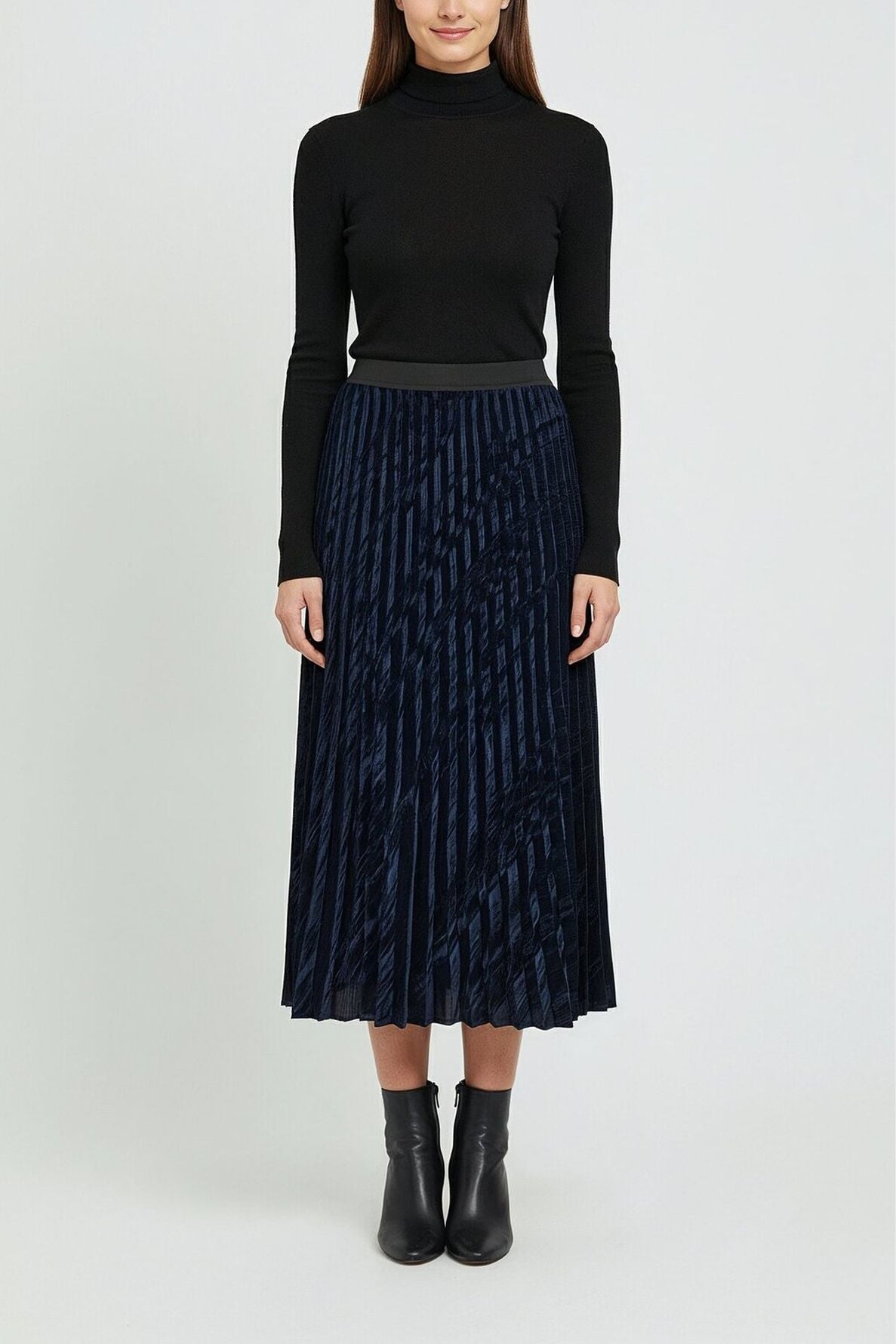 A woman stands facing forward, wearing a black turtleneck sweater, a navy blue jupe plissée midi skirt in velours frappé, and black ankle boots against a plain light background.
