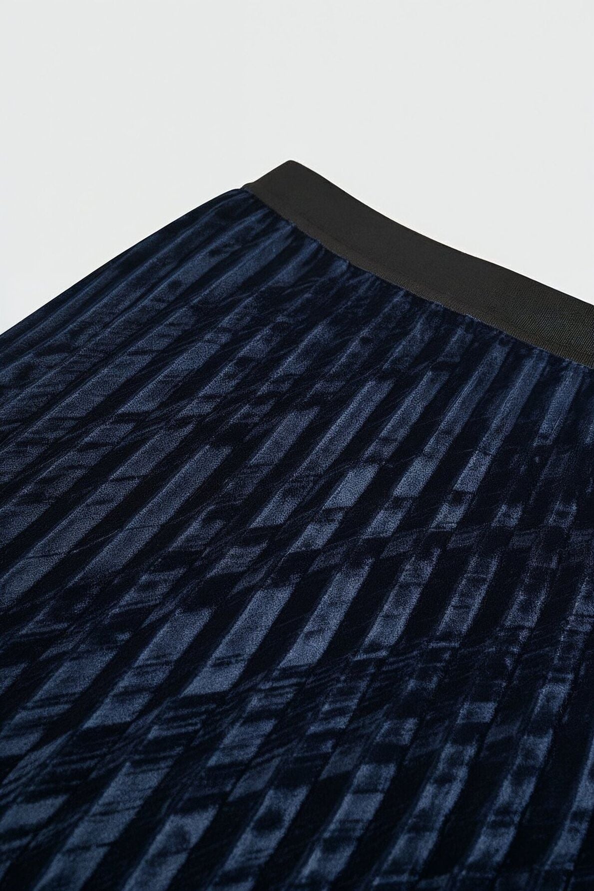 Close-up of a dark blue velours frappé pleated skirt with a black waistband, showing the textured fabric and subtle geometric pattern. This elegant jupe plissée falls at a midi length, highlighting both its luxurious material and refined style.