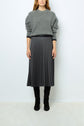 Woman wearing a loose grey sweater, a dark grey pleated skirt with subtle dots, and black knee-high boots, standing against a plain light background. Her face is partially out of frame.
