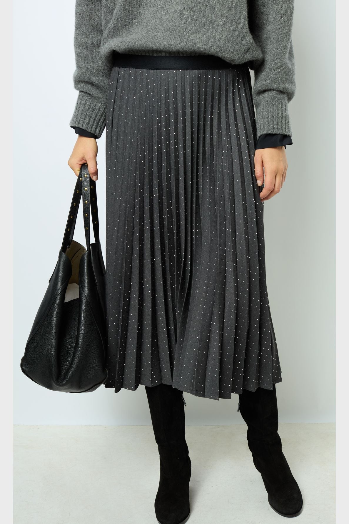 Person wearing a grey sweater, a black pleated skirt with small dots, black knee-high boots, and holding a large black handbag.