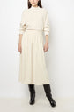 A person wearing a long-sleeve cream sweater tucked into a matching jupe midi velours, paired with knee-high black boots, stands against a plain white background.