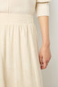 A person wearing a cream-colored ribbed sweater and a matching jupe hiver femme à fines côtes, shown from the waist down with one arm relaxed by their side.