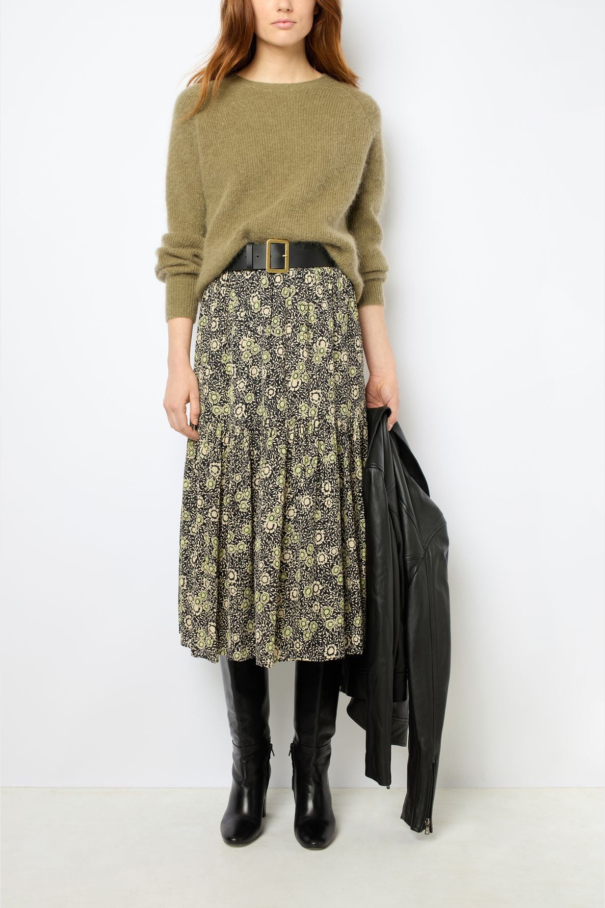 Woman wearing an olive sweater tucked into a floral midi skirt with a wide black belt, knee-high black boots, and holding a black leather jacket by her side.