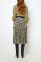 Woman standing with her back to the camera, wearing an olive sweater, a black floral midi skirt, a black belt, and knee-high black boots. Her long, wavy brown hair is loose. Background is plain white.