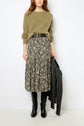Woman wearing an olive green sweater, black floral midi skirt with yellow flowers, black belt, and black knee-high boots. She holds a black leather jacket by her side.