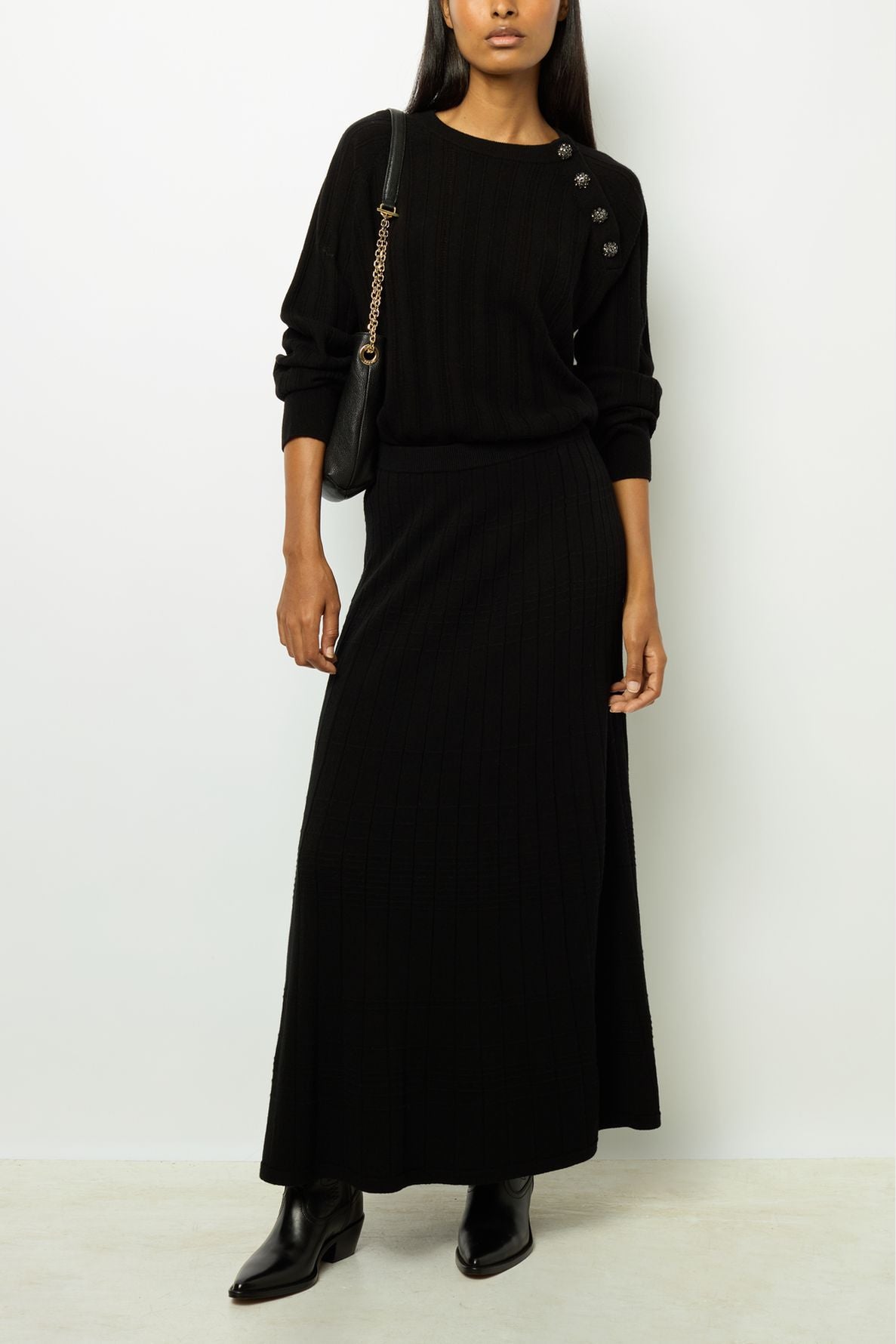 Woman wearing a black ribbed long-sleeve top with button details, a matching jupe midi en maille à côtes, black ankle boots, and a black shoulder bag with a gold chain strap, standing against a white wall.