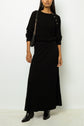 Woman wearing a black ribbed long-sleeve top with button details, a matching jupe midi en maille à côtes, black ankle boots, and a black shoulder bag with a gold chain strap, standing against a white wall.