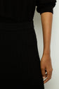 A person wearing a long-sleeved, ribbed black knit dress in maille à côtes stands against a light background, with their right arm relaxed at their side.