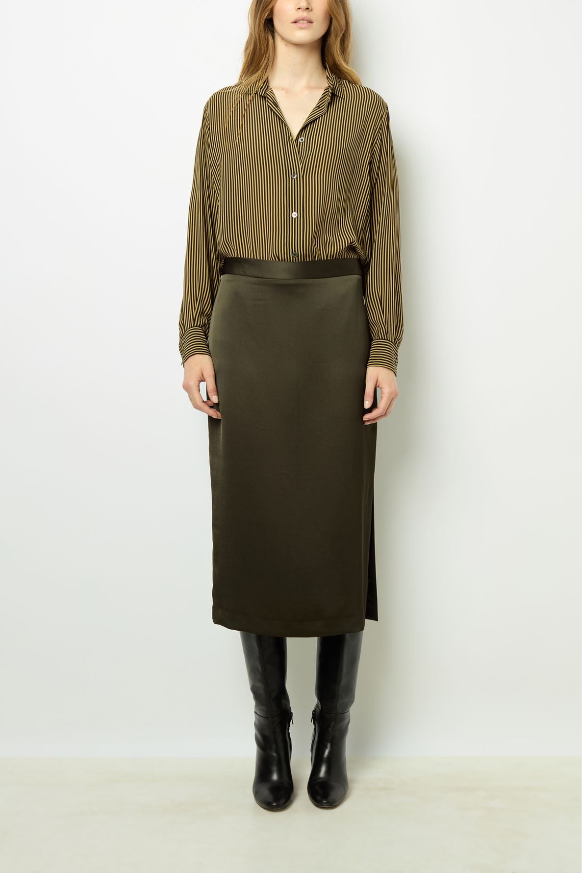 A woman stands against a white wall wearing a brown striped blouse, an olive green midi skirt, and knee-high black boots. Her hands are by her sides, and her hair is loose.