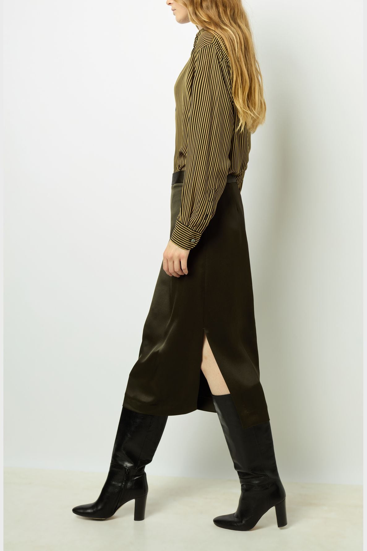 A person with long hair wears a striped blouse, a dark midi skirt with a side slit, and black heeled knee-high boots, walking against a plain white background.