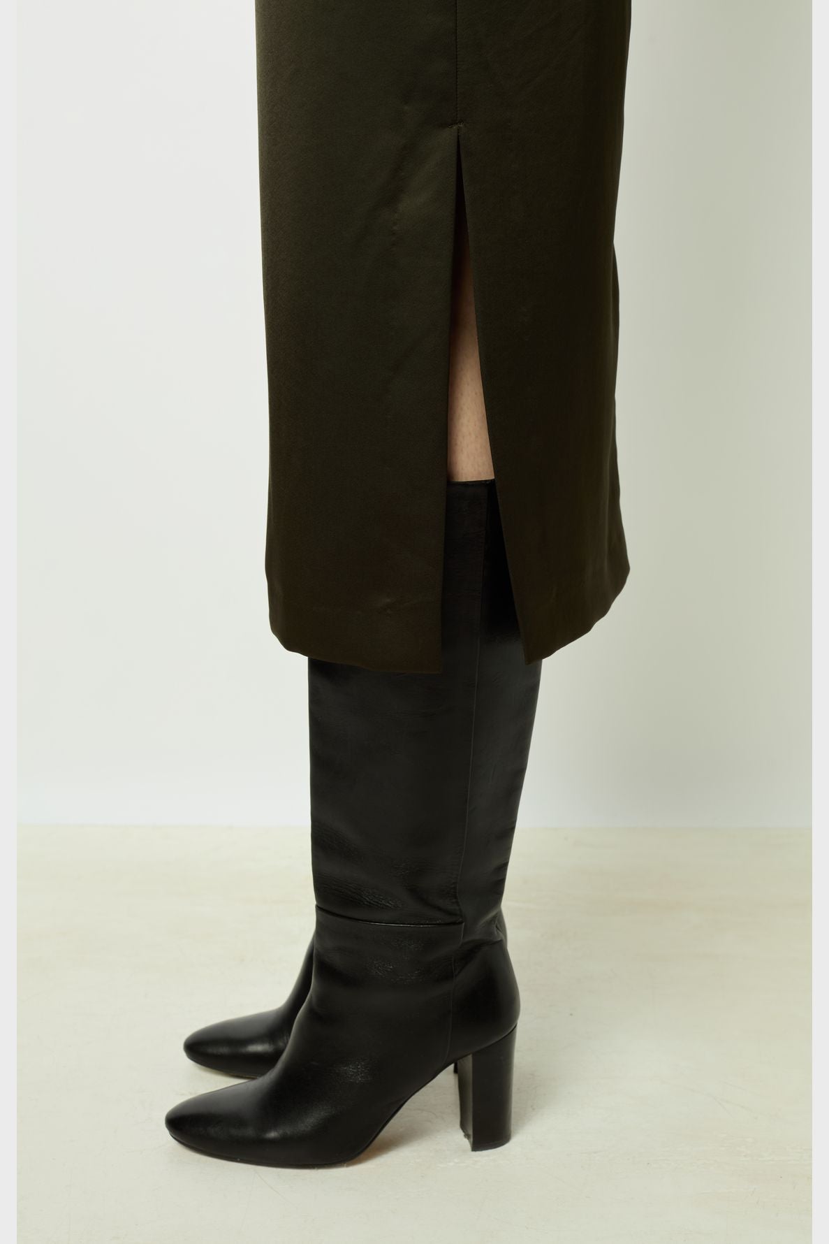 A person wearing a dark green skirt with a side slit and black heeled knee-high boots stands on a light-colored floor against a plain white background.