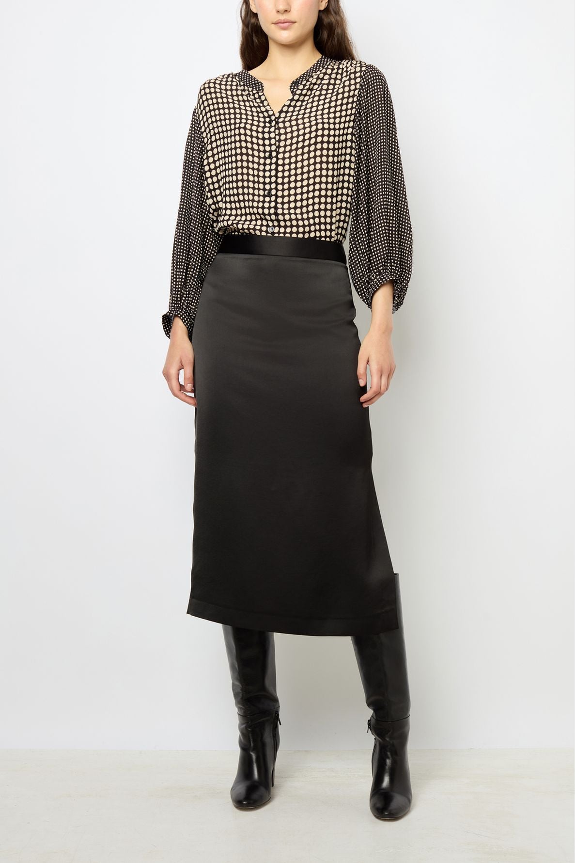 Woman wearing a patterned blouse with three-quarter sleeves, a jupe noire femme midi skirt, and black knee-high boots, standing against a plain white background.
