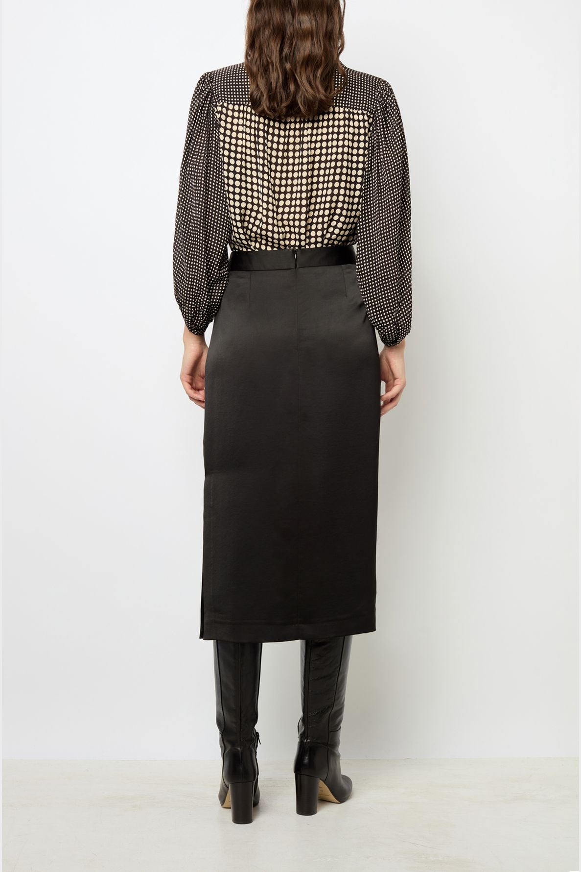 Woman standing with her back to the camera, wearing a black and white checkered blouse, a jupe noire femme chic—a high-waisted black midi skirt—and tall black boots, against a plain white background.