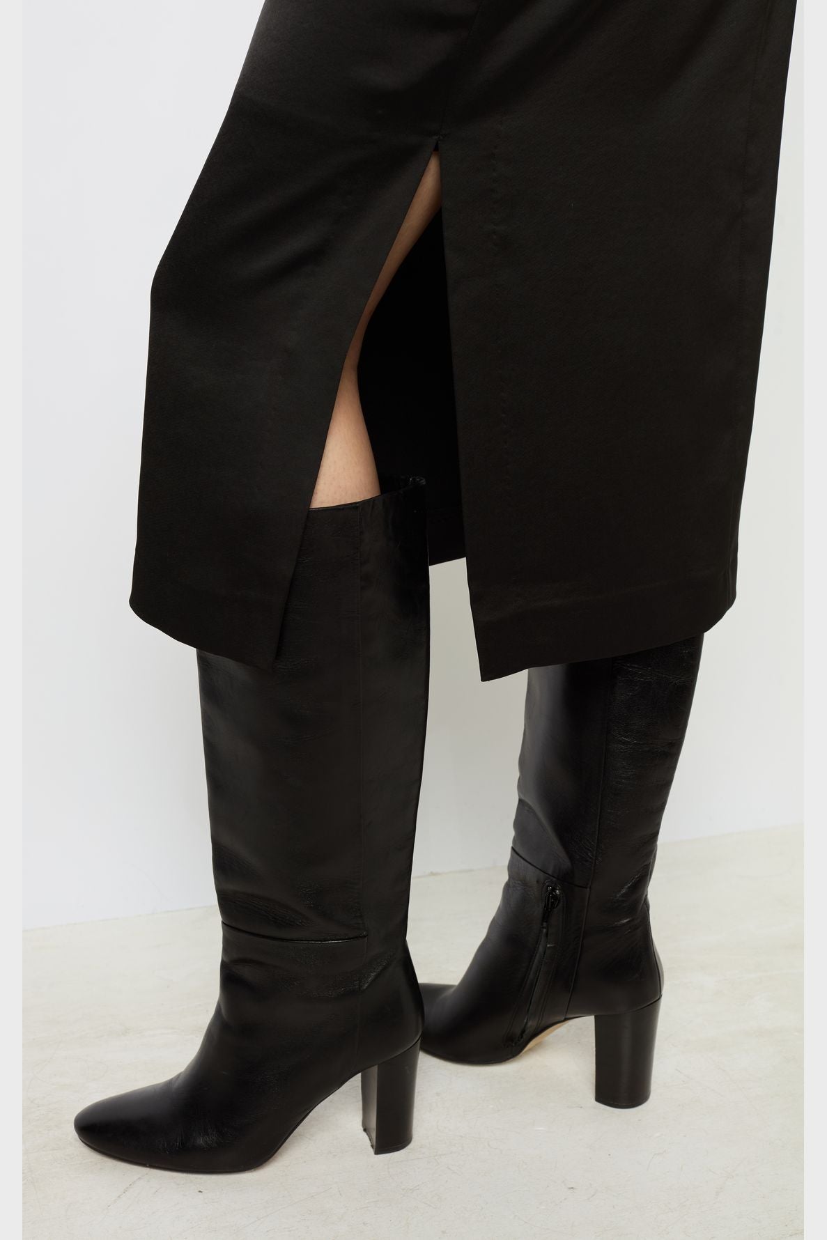 A person wearing a jupe noire femme with a side slit and knee-high black heeled boots stands on a light-colored floor, effortlessly showcasing a jupe chic style.