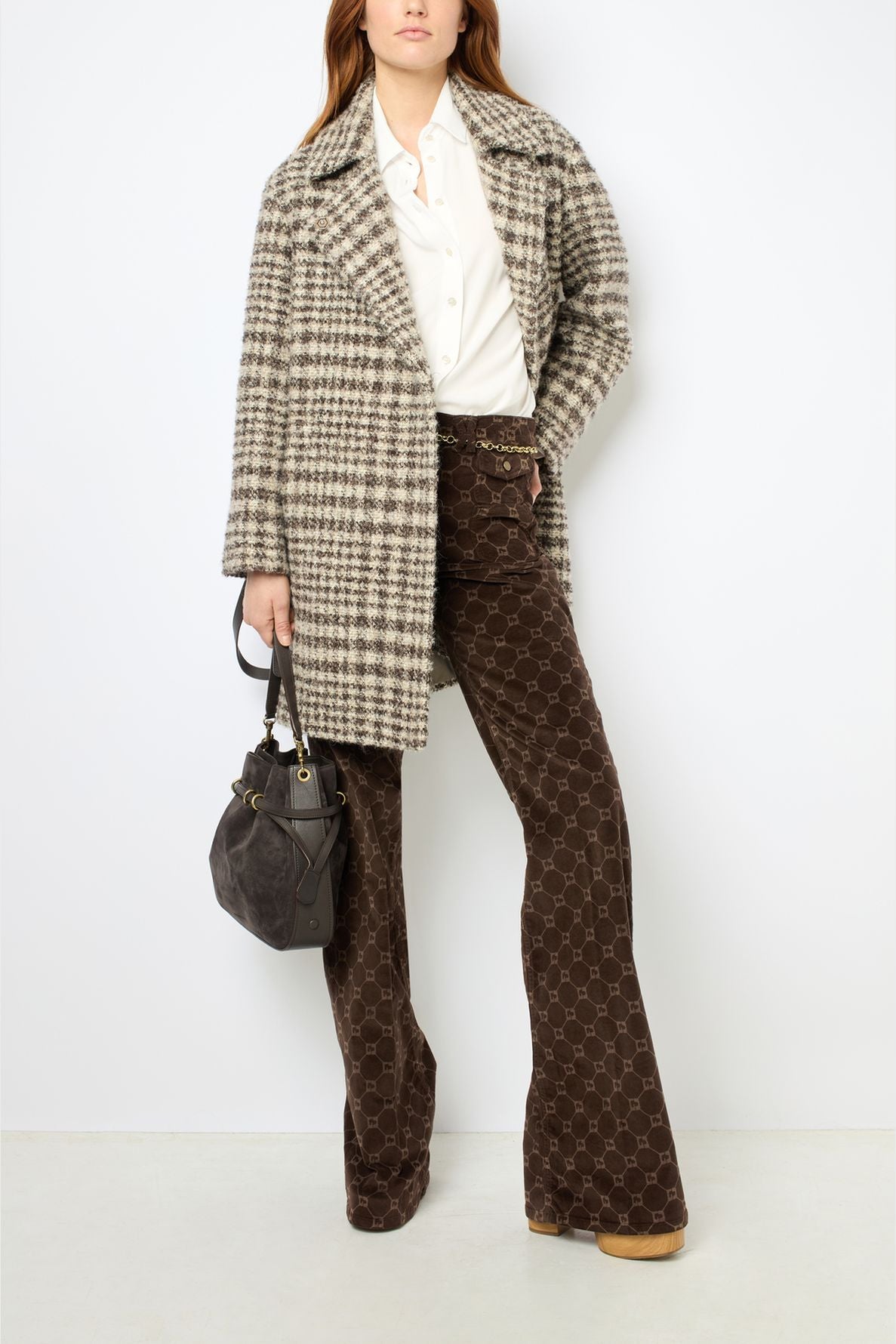 Woman wearing a manteau court à carreaux over a white shirt and patterned brown flared pants, holding a dark handbag, standing against a plain white background.