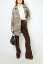 Woman wearing a manteau court à carreaux over a white shirt and patterned brown flared pants, holding a dark handbag, standing against a plain white background.
