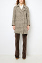 A person wearing a black and white houndstooth manteau laine alpaga over a white shirt and brown patterned pants stands against a plain white background.